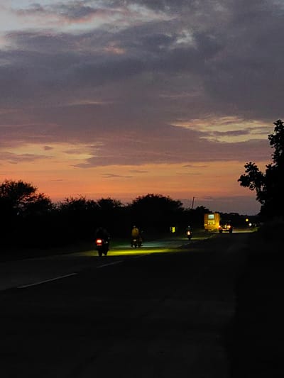 The image depicts a rural road at dusk, likely in or near Dattapur, India. The sky is a gradient of twilight colors, transitioning from a soft orange and pink near the horizon to a deep purple and gray with clouds higher up. Silhouetted trees and vegetation line the left side of the road.
Several vehicles are present on the asphalt road. In the foreground on the left, a motorcycle with its taillight illuminated is visible. Further down the road, another motorcycle is parked or moving slowly, its bright headlight casting a yellow glow on the road. To the right of this motorcycle, a person wearing a yellow jacket is standing or riding another motorcycle.
In the middle distance, a yellow truck or van with its headlights and taillights on is a prominent feature. A smaller vehicle, possibly a car, is behind it with its brake lights illuminated. Other distant lights suggest further traffic. The road itself has a white line marking its edge. The overall scene captures the dim light of evening on a journey or travel.