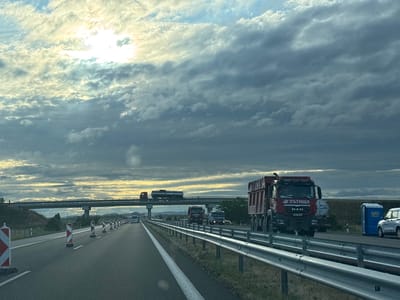 The image captures a view from a vehicle traveling on a highway in Bozduganovo, Bulgaria. The sky is filled with dramatic, cloudy formations with the sun breaking through in the upper left quadrant, casting a bright glow. The highway appears to be under construction or undergoing maintenance, with traffic cones and barriers lining the right side of the road. Several trucks are visible on the highway, including a large red dump truck with Cyrillic lettering and a logo that appears to be "ПЪТИЩА" (Pătishta), meaning "Roads" in Bulgarian. This truck is in the foreground on the right. Further down the highway, other vehicles, including a white bus and a smaller truck, are also present. An overpass stretches across the highway in the distance. On the right shoulder of the highway, a blue portable toilet and a white SUV are parked. The overall scene depicts a typical highway environment with traffic and roadwork, set against a backdrop of a dynamic sky.