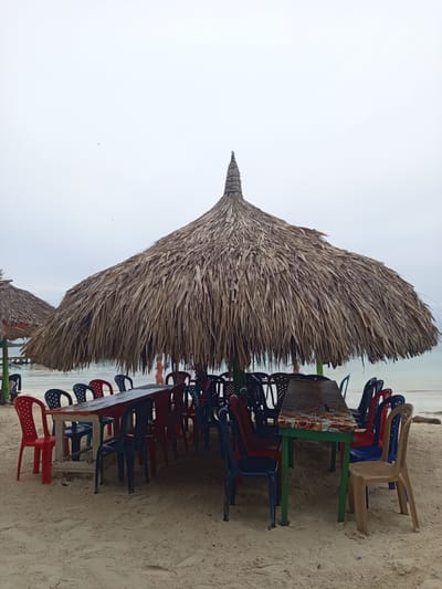 A thatched umbrella shades a long table and several plastic chairs on a sandy beach. The table is dark brown with a decorative edge, and it is supported by green legs. The chairs are a mix of red, blue, black, and tan. In the background, the ocean is visible, and beyond that, a wooden pier with some boats. A person wearing a pink bikini stands near the water. The sky is overcast. This scene appears to be a beachside dining area in a tropical location, possibly in a coastal city in Colombia given the mention of "Unknown, Colombia".