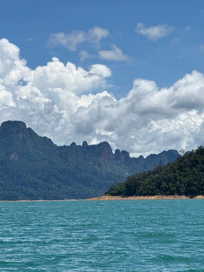 A wide-angle, eye-level shot captures a serene vista of the turquoise waters of Cheow Lan Lake in Khao Sok National Park, Ban Chiao Lan, Thailand. The lake occupies the foreground, its surface gently rippled by a light breeze. Beyond the water, a dramatic line of lush, green, jungle-covered karst mountains stretches across the mid-ground, their peaks sharp and jagged against the sky. A sandy shoreline is visible where the water meets the vegetation on a lower section of the mountains. The sky above is a vibrant blue, dotted with large, puffy white cumulus clouds. The scene is devoid of people and human activity, presenting a natural and tranquil landscape.