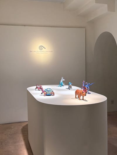 The image displays a modern art installation within a gallery. A curved, white display panel showcases several colorful, animal-like sculptures. The panel extends from the wall, under a recessed circular ceiling feature. The opposite wall bears the name "Jacobo & Maria Angeles," presumably the artists. The gallery has minimalist design, with neutral walls and flooring.