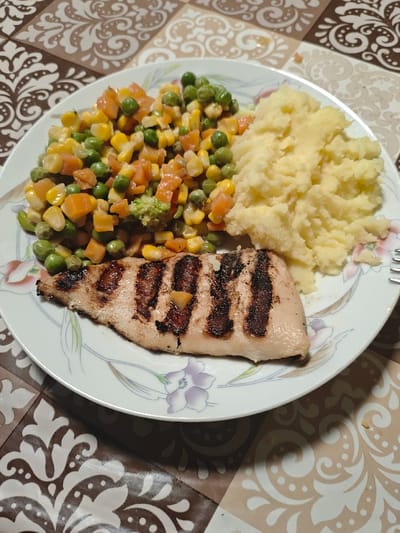 A white round plate, adorned with a floral pattern featuring lilac and pink flowers and green leaves, is positioned on a table covered by a tablecloth with an alternating brown and beige ornate pattern. The plate holds a meal consisting of three primary components: a grilled chicken fillet, a portion of mashed potatoes, and a mix of cooked vegetables. The chicken fillet, light brown with prominent dark brown grill marks, occupies the lower left section of the plate. To its right, a mound of pale yellow mashed potatoes is present. The upper half of the plate is filled with a medley of mixed vegetables, including orange diced carrots, green peas, yellow corn kernels, and some green broccoli florets. A silver-colored fork is partially visible on the right edge of the plate, resting on the tablecloth. The scene is located in the city of Opaka, Bulgaria.