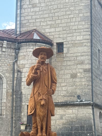 A wooden statue of a man wearing a wide-brimmed hat and a cape stands in front of a stone building. The man is holding a small object in his right hand and has a wolf statue at his side. The stone building appears to be a church or historical structure, with a peaked roof and arched windows visible. The sky above is partly cloudy. This statue is located in the city of Grande-Rivière Château, France.