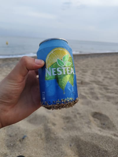 The image shows a hand holding a blue can of Nestea lemon iced tea at a beach in Pineda de Mar, Spain. The bottom of the can is covered in sand. The beach has light brown sand, and the sea is visible in the background. The sky is overcast. A yellow buoy is visible in the sea.