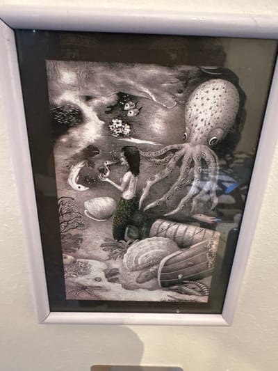The image displays a framed, predominantly grayscale artwork depicting an underwater scene, mounted on a light-colored wall in El Pinto, Spain. The illustration features a mermaid centrally positioned, possessing dark hair and a scaly, dark green tail. She holds a small, light-colored, duck-like creature in her hands, with small bubbles visible around it. To the upper right, a large octopus extends multiple tentacles across the scene.
The underwater environment includes a detailed seafloor with various marine elements. Prominent objects on the seafloor comprise large seashells, including a ribbed oyster shell in the lower right, and broken wooden structures resembling barrels or boat fragments. Marine life observed beyond the main subjects includes a long, light-colored fish near the mermaid's left, two striped fish in the upper-center, and a smaller, elongated fish in the mid-right. A starfish is visible on the seafloor in the lower-left. Coral-like formations and aquatic plants are distributed throughout the scene, with patterns suggestive of light filtering through water present in the upper-left quadrant. The artwork is enclosed within a simple white frame, and reflections are discernible on its glass surface.