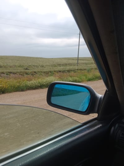 The image shows a view from inside a car, looking out towards a rural landscape. A side-view mirror with a blue tint reflects a road and sky. Beyond the road, there are fields of green and brown vegetation, with a few small wildflowers. A utility pole is visible in the distance against a cloudy sky. The interior of the car is partially visible, including the door panel and window. The location is identified as Aleksandrovka, Kazakhstan.