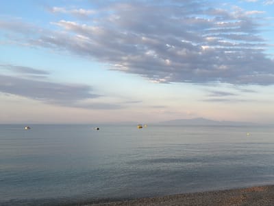 The image depicts a serene seascape at Νέα Φλογητά, Greece, likely during a sunset or sunrise. The foreground features a pebble beach, transitioning into a calm, blue sea that stretches to the horizon. Several small boats are visible on the water, some appearing to be moored. In the distance, a faint outline of mountains or landmasses can be seen against a sky filled with soft, streaky clouds in shades of blue, white, and pale pink. The overall atmosphere is peaceful and tranquil.