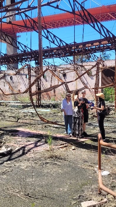 The scene depicts an outdoor environment at a derelict industrial site in Zárate, Argentina, under a clear blue sky. The ground is uneven, composed of dark soil, scattered debris, and sparse vegetation, with shadows cast across it. In the midground, four individuals are standing. From left to right: an adult female in a light blue collared shirt and blue jeans, wearing sunglasses; a young adult female in a black and white patterned long skirt and a dark top; a young adult male in a black t-shirt, dark shorts, and a black baseball cap, holding a dark object and a red can; and an adult female in dark clothing and a wide-brimmed light-colored hat, facing the group while holding a small object and gesturing. The individuals appear to be interacting or observing. The background is dominated by the extensively rusted steel framework and concrete remnants of a large, partially collapsed industrial building. Above this structure, a segment of a large, red-painted suspension bridge, identifiable as part of the Zárate-Brazo Largo Bridge complex, is visible against the clear sky, along with associated support columns and overhead electrical lines.