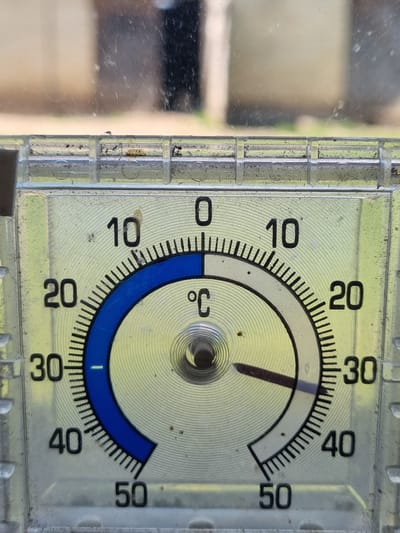 This is a close-up, high-angle shot of a thermometer. The thermometer has a clear plastic casing and a dial with markings from 0 to 50 degrees Celsius, indicated by black numbers and tick marks. A thin red line within the thermometer indicates the temperature. The temperature shown is between 20 and 30 degrees Celsius, appearing to be around 23 degrees. There are blue markings on the dial from 10 to 20 and from 30 to 40 degrees Celsius. The thermometer is dirty and has some smudges on the surface. The background is blurred and appears to be outdoors, with some greenery visible. The overall impression is of an outdoor thermometer measuring a warm day. The visible text is "°C", and the numbers 0, 10, 20, 30, 40, and 50. The location is Žygaičiai, Lithuania, suggesting the weather is likely warm or temperate for that region.