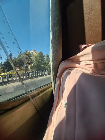 The image depicts an outdoor urban scene in Ashrafia, Syria, viewed from inside a vehicle. A transparent ruler, marked with metric measurements including numerals like 87, 88, 89, and 90, is positioned vertically against the vehicle's window, partially obscuring the external view.

Outside the window, a clear blue sky fills the upper portion of the frame. Below the sky, a multi-story building featuring a darker rooftop structure is visible, partially obscured by a line of green trees. In the mid-ground, a road with multiple lanes is separated by a series of grey road barriers. A yellow vehicle is visible on the far side of these barriers.

On the right side of the image, within the vehicle's interior, a light pink garment, characterized by visible stitching and a white drawstring, is prominently displayed. Sunlight casts distinct shadows across the fabric. The vehicle's interior framing the window appears to consist of dark brown and lighter, textured paneling.