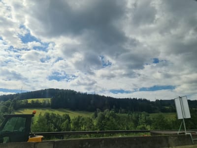 The image shows a roadside view in Spital am Semmering, Austria, featuring a cloudy sky over a lush green landscape. In the foreground, a portion of a yellow construction vehicle with an orange flashing light is visible on the left, positioned behind a concrete barrier. A black metal railing runs horizontally across the middle ground. Beyond the railing, a sloping green meadow is dotted with trees, leading up to a dense forest that covers the hillside. In the distance, a power transmission tower is visible on the crest of the hill. A blank white sign on a green pole stands on the right side of the road.