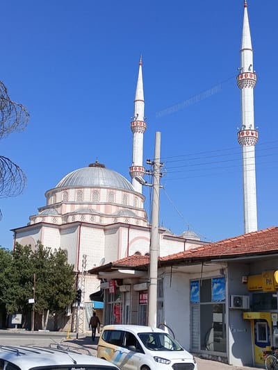 The image shows a mosque with a large dome and two minarets against a clear blue sky. In the foreground, there are several buildings with tiled roofs, including what appears to be a PTT (Post, Telegraph, Telephone) office. A white car is parked on the street, and a person is walking away from the camera on the sidewalk. Trees and a utility pole with electrical wires are also visible. The mosque exhibits traditional Islamic architecture, with decorative elements on the minarets and windows. The overall scene depicts a street view in a town or city, likely in Turkey, given the presence of the PTT office and the architectural style.