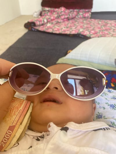 In Arbagish, Uzbekistan, a close-up shot shows a baby's face partially obscured by large, white-framed sunglasses with purple lenses. A hand holds the sunglasses, and a stack of Uzbekistani 2000 Som banknotes is placed near the baby's cheek. The background includes blurred floral-patterned fabric and bedding.