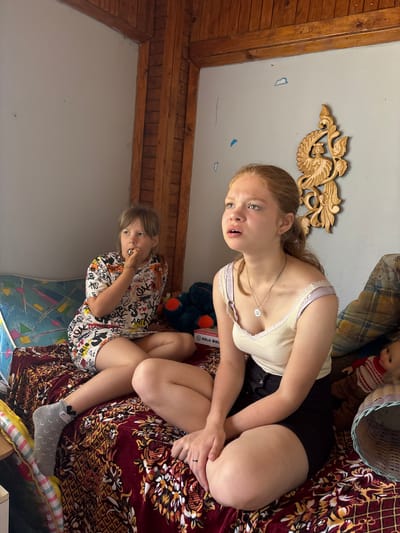 The image depicts an indoor scene with two young girls seated on a patterned blanket spread across a low surface, possibly a bed or sofa. The girl on the left, with blonde hair and bangs, wears a white dress adorned with colorful graffiti-style text and illustrations. She is holding something to her mouth and appears to be eating or putting something in her mouth. Her left leg is crossed over her right, and she wears a grey sock with a black design on her left foot. The girl on the right, also with blonde hair tied back in a ponytail, is wearing a cream-colored tank top with a lace trim and black shorts. She is looking upwards and to the left with an open mouth, as if speaking or reacting to something. Her legs are crossed, and she is wearing a necklace with a circular pendant.
The background features a wall painted a light gray, with wooden paneling and beams in the upper portion of the image. A carved wooden ornament, possibly depicting a bird-like creature with a human face, is mounted on the wall above and to the right of the girl on the right. To the right of this ornament, a pillow with a plaid pattern is visible, and next to it, a doll and a woven basket. To the left of the girls, a bolster or pillow with a geometric pattern is partially visible. The overall environment suggests a rustic or simple living space. The presence of wooden elements and the style of the furniture and wall decorations hint at a location in a more rural or traditional setting. The question asks for the location in the city of Аряж, Russia, but there is no visual information in the image that can confirm or deny this specific location.
