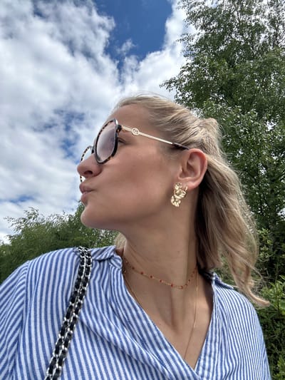 A woman in a blue and white striped shirt stands outdoors. She wears gold earrings and sunglasses. A gold chain necklace with red beads and a black and silver purse strap are visible. The background includes trees, blue sky, and white clouds. No specific evidence confirms the location is Saint-Priest-en-Jarez, France.