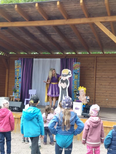The image shows an outdoor stage in a wooded area. A woman with blond hair, wearing a purple dress with a gold cape and belt, stands on the stage, gesturing towards the audience. Behind her, a backdrop includes purple and white curtains, along with comic-style banners with the words "WELCOME" and "HEROE". There are cardboard cutouts of cartoon characters on the stage as well. In front of the stage, a group of children are standing and watching the performance. The date and time are displayed in the lower right corner: "Hradec Králové 8 май 2026 г. в 14:19 ч." Based on the name "Hradec Králové" in the OCR, the location might be near the city with the same name in Czechia. However, Chlumec nad Cidlinou is closer to Hradec Králové than other cities. Further details from the original image and possibly associated location details would be needed to confirm the exact location.