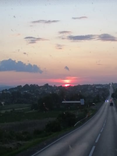 The image depicts a dusk scene from a moving vehicle on a road leading towards a distant town. The sky is a gradient of pale blue and pink, with scattered clouds. The sun is low on the horizon, casting a reddish glow. A town or village is visible in the middle ground, nestled among trees and rolling hills. The road in the foreground is paved and marked with white lines, with a few vehicles visible in the distance. The overall atmosphere is serene, suggesting a journey concluding as night falls. The location is identified as Dărmănești, Romania.