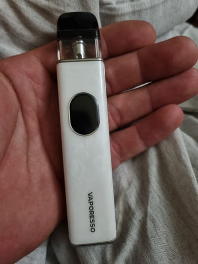 A hand is holding a white Vaporesso vape pen against a gray, textured fabric. The vape pen is rectangular with rounded edges. On its surface, the word "VAPORESSO" is printed in black text. There is also a black, oval-shaped button or screen on the vape pen, located towards the left side. The hand is resting on the fabric, and the fingers are slightly curled around the vape pen. The lighting suggests it is indoors, and no specific time of day or weather conditions can be determined. The scene is simple, focusing on the object and the hand. The provided location context is Chuhuiv, Ukraine, but this information cannot be visually confirmed from the image itself.