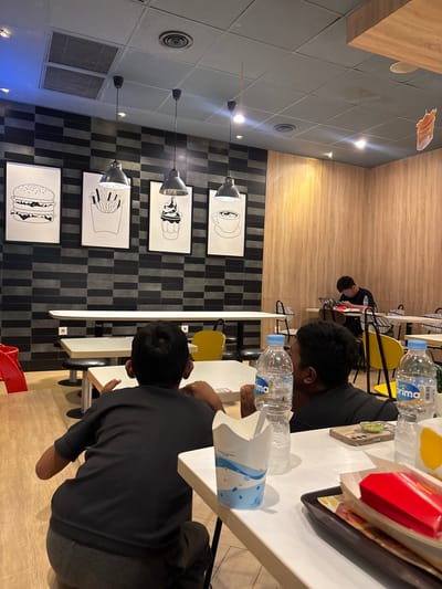 The scene is inside a McDonald's restaurant, presumably in Bencongan Indah, Indonesia. Three people are seated at tables. At the closest table, a person is seated, wearing a dark-colored top and grey pants. On the table are several McDonald's items including a drink cup, a red box, and bottled water. Further away, a person wearing a dark shirt sits alone at another table. The restaurant's interior includes a tiled ceiling, hanging lights, and decorative posters of McDonald's menu items on the wall. The tables are white with various chair styles.