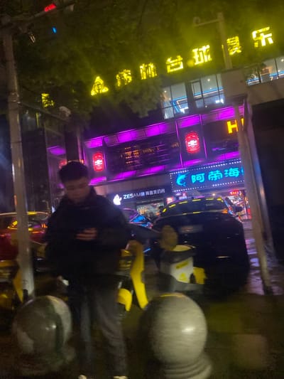 The image depicts a nighttime urban street scene in 天宫殿街道, China, illuminated by artificial lighting. In the immediate foreground, several grey, spherical stone bollards are visible, partially blurred. To the left, a vertical metal pole extends upwards. In the center foreground, a male figure stands, wearing a dark puffer jacket and dark trousers, looking downwards. His face is indistinct due to blur and low light. Directly behind him and to his right are yellow and grey shared mobility devices.
The ground surface, appearing wet, reflects various lights. In the mid-ground, a dark-colored sedan is visible, parked or moving slowly, facing right, with other vehicles partially obscured further back. The background features a multi-story commercial building. Its upper facade displays a large yellow neon sign, partially obscured by green foliage, reading "金街台球娱乐" (Golden Street Billiards Entertainment). Below this, the building's mid-section is prominently lit with purple neon strips. At a lower level, a blue neon sign features a stylized fish or dolphin icon alongside Chinese characters "阿帝海!" (Ah Di Hai!). To the left of this sign, a white sign reads "ZESAM 赛膳美药房" (ZESAM Saisanmei Pharmacy).