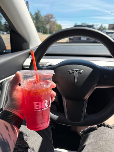 A person's hand holds a clear plastic cup filled with a pink-colored beverage and ice, with a striped straw inserted. The cup has a Baskin-Robbins logo visible on its side. The person is wearing a black mesh watch on their left wrist and is seated in a car, indicated by the presence of a car steering wheel with a Tesla logo in the center. The background shows a slightly out-of-focus view of a street with trees and other vehicles, suggesting an outdoor setting. The image was taken in Buffalo Grove, United States of America.