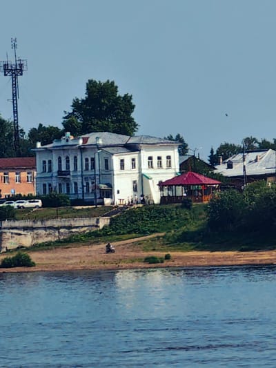 The image shows a waterfront view of Yeniseysk, Russia, with several buildings and natural elements. In the background, a tall communications tower stands against a clear blue sky. A large, light-colored, multi-story building with many windows and ornate architectural details is prominently featured. To its left, a smaller, reddish-brown building is visible. In front of the buildings, a red-roofed gazebo sits on a grassy slope. A body of water, likely a river, occupies the foreground, with reflections of the sky and surrounding scenery on its surface. The riverbank is lined with sand and vegetation. On the bank, a person is riding a motorcycle. Several white cars are parked near the buildings, suggesting a public area or road. The overall scene depicts a tranquil day in a riverside town.