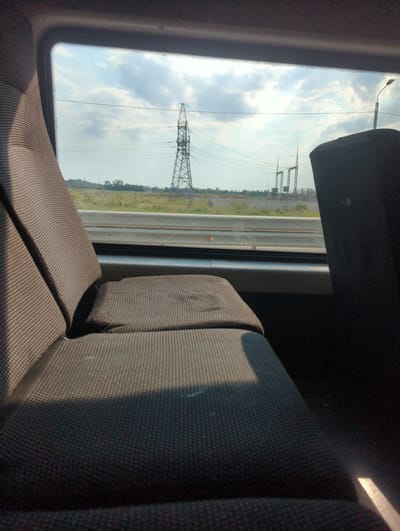 The image shows the interior of a train, looking out the window. The seats in the foreground are upholstered in a dark, patterned fabric. Outside the window, a landscape with a power line tower and a substation can be seen under a cloudy sky. The location is given as Укромновское сельское поселение, Ukraine.
