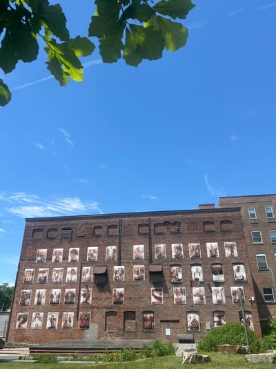 This image depicts a large, brick building adorned with a grid of framed portraits. The building is located in the Town of New Windsor, United States of America. The portraits appear to be of individuals, possibly a historical exhibition or a community art project. The building itself has a weathered brick facade with visible windows and some architectural details. In the foreground, there is a grassy area with some rocks and what appears to be a seating area or stage structure. The sky is a clear blue with a few wispy clouds, and the tops of some green leaves are visible in the upper left corner, suggesting it's a bright, sunny day. The overall scene is an outdoor public space, likely a park or plaza adjacent to the building, showcasing art or historical imagery.