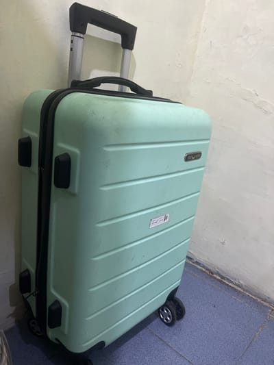 This is a media file featuring a light green, hard-shell suitcase with a textured surface. The suitcase has a telescoping handle with a black grip and silver metal extensions, as well as two black wheels visible at the bottom right. There are also two black handles on the side of the suitcase. A small white sticker with some writing is attached to the top surface. Another label, also with text, is visible near the top of the suitcase. The suitcase is positioned on a floor that appears to be tiled in a blueish-grey color, next to a wall that is painted white and has a textured, possibly stuccoed, finish. The lighting suggests an indoor setting, possibly a room or a storage area. There is no apparent activity or event taking place; the suitcase appears to be stationary. The scene is simple and functional, focusing on the object itself. The visible text on the labels is not clearly legible in the provided image. The location context of Anggaswangi, Indonesia, is noted, but there are no specific visual cues in the image to confirm this location.