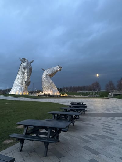 The image displays an outdoor scene at The Helix park in Falkirk, United Kingdom, at twilight or under heavily overcast conditions. The primary subjects are two monumental, highly detailed metallic horse head sculptures, known as The Kelpies, positioned centrally in the background and illuminated from below by warm-toned lights. The left sculpture depicts a horse's head facing forward, while the right sculpture shows a horse's head raised with an open mouth. Both are constructed from numerous faceted metal plates. In the foreground and midground, a linear arrangement of approximately six dark-colored wooden picnic tables with attached benches occupies a gray patterned paved walkway. To the left of the walkway is a maintained green grass area. The background features a line of bare deciduous trees, a street lamp emitting light, power lines, and elements of distant infrastructure including a bridge and low walls. The sky is dark and predominantly covered by dense, grey clouds. No individuals, actions, or interactions are present in the scene.