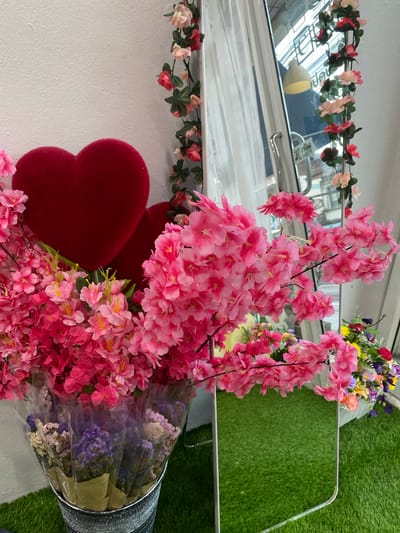 In Khok Kloi, Thailand, a close-up shot captures a decorative arrangement featuring two plush red hearts and an abundance of vibrant pink artificial cherry blossoms. The flowers are arranged in a tall, galvanized metal bucket, with smaller dried flowers in shades of purple and cream at the base, wrapped in paper and clear plastic. To the right of the bucket, a rectangular mirror with a silver frame reflects the scene, showing artificial greenery and a colorful bouquet of flowers in a white pot. A string of pink and red artificial roses with green leaves is draped vertically along the right edge of the image, bordering a white wall. The background, seen through the mirror, suggests an indoor commercial space, possibly a cafe or shop, with a reflective ceiling and glass doors. The overall setting is well-lit and appears to be a commercial establishment, likely a florist or a shop decorated for a special occasion, possibly Valentine's Day, given the heart motif.
