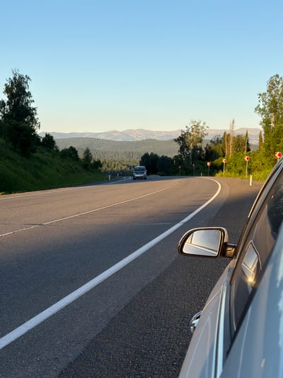 This is an eye-level, medium shot of a paved road with a white line marking the lane. The road curves to the left, bordered by a grassy embankment on the left and trees on the right. A silver car is parked on the right side of the road, its side and side mirror visible. In the distance, a white van is driving away from the camera on the road. The background consists of a dense forest leading to rolling hills and mountains under a clear blue sky. The scene appears to be set in the early evening, with warm sunlight casting long shadows. The location is a rural road outside the city of Kaz, Russia.