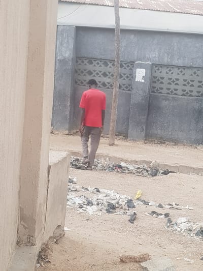 An adult male wearing a red shirt and gray pants is urinating beside a gray-colored building in Dutsinma A, Nigeria. The building is made of concrete blocks with a decorative geometric pattern and a small white sticker affixed to its surface. A narrow channel filled with discarded trash and debris runs between the building and an adjacent sandy area. A broken tree branch lies on the ground. The surrounding area appears to be dusty and unpaved. The lower portion of a concrete structure is visible in the foreground.