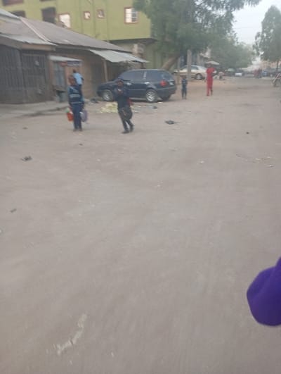 A street scene reveals a dusty road with scattered debris. Several people are visible, including two children carrying items. A dark-colored car is parked near buildings with corrugated roofs. Another person in a red outfit stands further down the road. Trees are scattered along the background. The overall atmosphere appears somewhat rundown and dusty. A purple object is visible in the lower right corner.