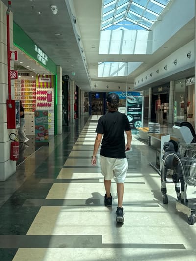 This media file shows a person walking down a brightly lit indoor shopping mall corridor in Carini, Italy. The person is wearing a black t-shirt, white shorts, and a baseball cap, and appears to be pushing a shopping cart. Sunlight streams in from a large glass ceiling, casting distinct shadows on the marble floor. The corridor is lined with shops, some of which have displays visible. Text in the image includes shop names and signs such as "LA CASUR IS", "POWER", "TENDENZE", "SUMMER", "CRAZY DUMPLING", "W.C.", "GIFT OGATORE", "AV COSTRUZIONI", and "CAMOMILL". The overall impression is a typical day of shopping in a modern mall. There are no obvious emotional cues or indications of specific weather conditions beyond the bright interior lighting. The presence of a fire extinguisher and signs related to emergency equipment suggest standard safety measures are in place.