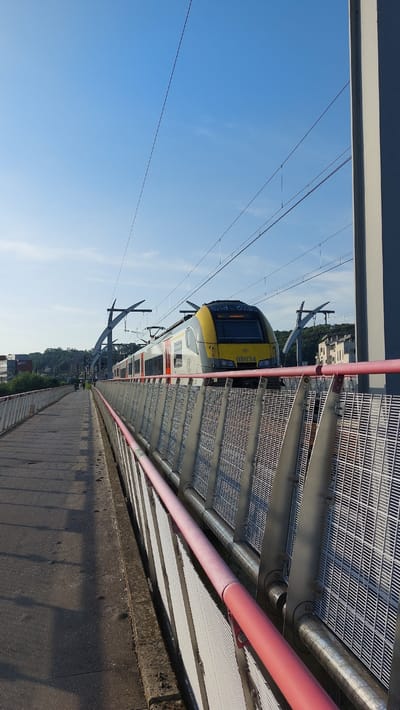 The image shows a train at a station on a bright, sunny day. The train is modern and yellow and silver, with the number "08034" visible on the front. It is positioned on tracks that are elevated above the ground, with a metal platform and overhead wires visible. The scene appears to be outdoors, with a clear blue sky and some distant greenery and buildings. The perspective is from the platform looking down the length of the train and the track. The lighting suggests it is daytime, possibly morning or afternoon given the bright sunshine and clear sky. The overall impression is of a functional and modern railway station. There is text on the train that reads "First Class" and "08034". The location is Namur, Belgium.