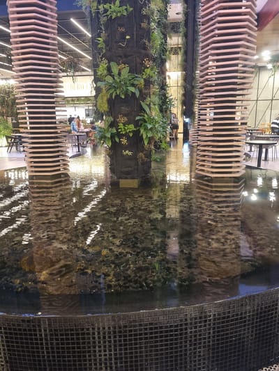 The image shows a tranquil indoor scene featuring a shallow body of water with a visible rocky bottom, possibly a fountain or water feature, in the foreground. The water reflects the ambient light and surrounding structures. Two large, hexagonal, wooden pillars with a layered, architectural design stand prominently. One is on the left and the other on the right, with a central, darker, cylindrical structure covered in lush green plants, forming a vertical garden.

In the background, several people are seated at tables in what appears to be a food court or restaurant area within a commercial space. The ceiling above shows a modern lighting system. The overall atmosphere is calm and contemporary, with a focus on natural elements integrated into the urban environment. The location is the Castelo da Coca-Cola in Araçariguama, Brazil, which is a historical landmark and a popular tourist attraction that has been repurposed.
