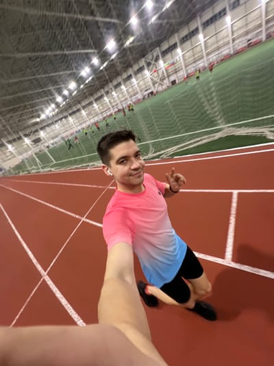 A man poses indoors on a red track, likely in an indoor sports facility with netting on the left. He wears a gradient pink and blue shirt, black shorts, and black shoes. He holds up his right hand with fingers slightly curled. The ceiling of the facility is visible in the background with overhead lights and people in the distance. The image is taken from a low angle.