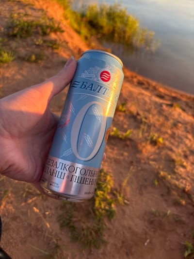 A hand holds a light blue aluminum can of "Baltika 0" non-alcoholic wheat beer near a body of water with a sandy bank. The can has "BALTIKA" in large blue letters and a prominent white "0" symbol. The background shows a sandy embankment with sparse vegetation, illuminated by the warm light of sunset. The water reflects the sky and surrounding greenery. The scene is set in the vicinity of Sapunov Bor, Russia, a location known for its natural landscapes.