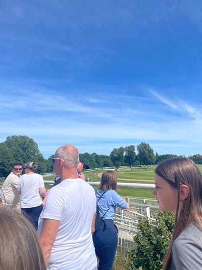 This media file captures an outdoor scene at what appears to be a racetrack in Wysoka, Poland, under a bright, sunny sky. The image is taken from an elevated perspective, looking down and across a green track area bordered by white railings.
Several people are gathered, observing an event. In the foreground, prominently positioned, is a balding man with short grey hair and sunglasses, wearing a white t-shirt, looking forward. To his left and slightly behind, another person with hair tied back also wears a white t-shirt. Behind them, a woman with medium-length brown hair, wearing a light blue long-sleeved top and dark blue pants, leans on a metal fence, also looking towards the track. In the upper right corner, a young woman with long brown hair, wearing a grey t-shirt, is visible from the side, looking downwards. Other partial figures and heads are visible throughout the crowd.
The setting features a wide expanse of green grass, indicative of a racing track or course, with white fences and railings defining its edges. Beyond the track, a line of lush green trees stretches across the horizon under a clear, bright blue sky with wispy white clouds. The strong sunlight and lack of heavy shadows suggest it is midday or early afternoon. No visible text is present in the image. The general atmosphere suggests a leisurely event or gathering on a pleasant day.