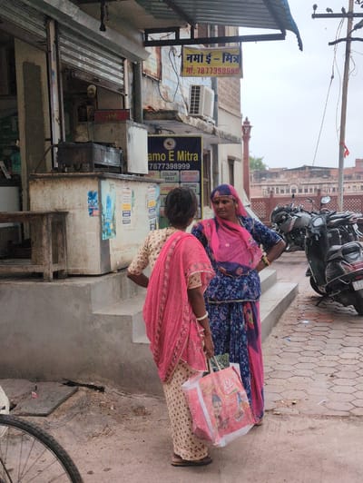 This is a medium-shot, eye-level, outdoor photograph taken in Jodhpur, India. The scene depicts two women interacting on a paved walkway in front of a building. The woman on the right, wearing a blue floral-print sari and a pink headscarf, is facing left and speaking to the woman on the left. The woman on the left, her back mostly to the camera, wears a cream-colored sari with a pink border and a pink scarf draped over her shoulders. She is holding a pink and white reusable shopping bag featuring an image of a young girl and the word "Masala." Behind the women, a row of motorcycles is parked along the walkway. The building behind them appears to be a shop with a metal roll-up door. A sign above the shop reads "namo e-mitra" in Hindi and English, with a phone number "9877398999." Another sign below that advertises services such as "account," "work," and "driving." The ground is paved with hexagonal tiles. A portion of a bicycle wheel is visible in the lower-left corner of the frame. The sky is overcast.