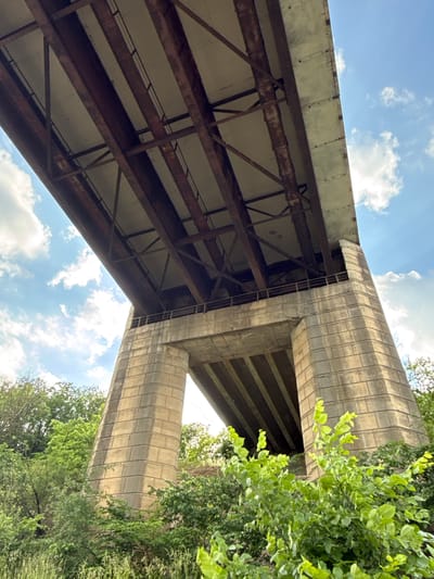 An underside view of a large concrete and steel bridge is captured in the image, with a bright blue sky dotted with white clouds visible above. The bridge structure is complex, featuring exposed steel girders and beams supporting a concrete deck. The concrete piers supporting the bridge are massive and appear to be constructed from large, rectangular blocks. Lush green foliage and trees surround the base of the bridge, suggesting a natural or semi-natural environment. The lighting indicates it is daytime, likely with sunlight casting shadows. The image does not contain any people or specific actions. This bridge is located in the city of Volkhov, Russia.