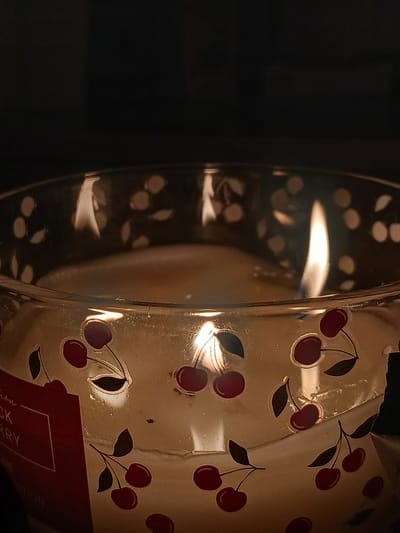 A lit candle in a glass jar with a cherry pattern is in focus. The candle is a light cream color and appears to be partially melted. The glass jar has a cut-out pattern of cherries and leaves. The background is dark and out of focus. There is a red label on the left side of the jar that says "Edition BLACK CHERRY HOME EDITION". There are no people or animals visible in the image. The location of this image in Rukla, Lithuania, is not discernible from the visual information provided.