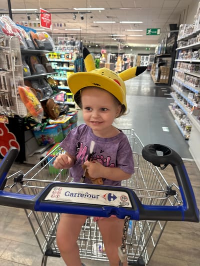 A young child, wearing a yellow Pikachu hat and a purple t-shirt, sits in a Carrefour shopping cart in a brightly lit supermarket aisle. The child is smiling and holding a toy. Shelves stocked with various merchandise, including bags and toys, are visible in the background. The shopping cart has the Carrefour logo on the handle. The scene suggests a shopping trip in a supermarket in Berchem-Sainte-Agathe - Sint-Agatha-Berchem, Belgium.