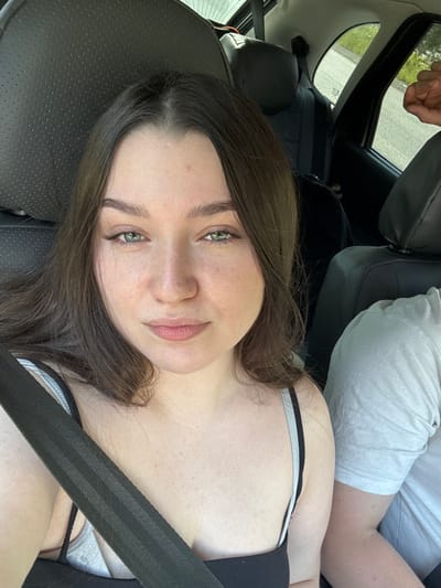 The image shows a young woman with long brown hair and green eyes, wearing a black tank top and a grey bra, with a seatbelt across her chest. She is seated in the front passenger seat of a car, looking directly at the camera. The interior of the car is visible, including the headrest of the driver's seat and a portion of the passenger seat. The window on the right shows a blurry view of trees and a road, suggesting the car is in motion. Another person's arm, wearing a light-colored t-shirt, is visible on the right side of the frame. The scene is lit by natural daylight. There is no indication of the city of Pesterevo, Russia.
