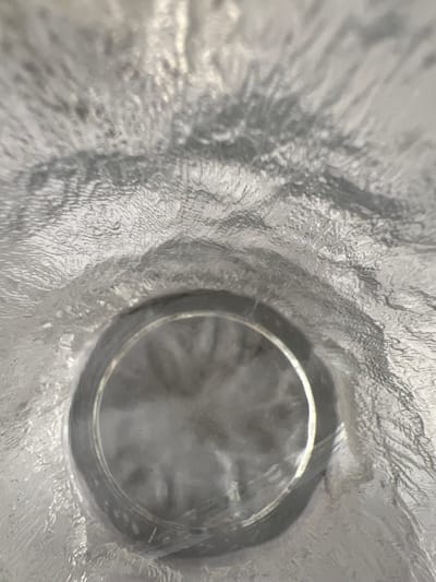 This is a close-up, abstract shot looking down into a clear, textured cylindrical object. The interior walls of the object are highly textured, with a pattern that resembles ice crystals or rough glass. The texture creates a rippling, distorted effect on the light passing through. In the center of the frame, there is a circular opening, through which one can see a blurry, diffused light. The lighting suggests that the object might be made of glass or plastic and that it is either filled with a clear liquid or is empty and catching ambient light. The overall impression is one of looking into a frozen or transparent cavity. There is no visible text. The location context of Ornek, Kyrgyzstan, does not offer specific visual cues within this abstract image.