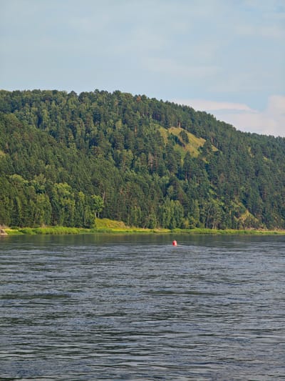 The image depicts a wide river flowing in front of a densely forested hill under a bright, partly cloudy sky. A red buoy floats in the middle of the river. The hill, covered in various shades of green trees, slopes upwards from the riverbank, with some patches of lighter green vegetation or exposed earth visible on its side. The river surface is rippled, reflecting the colors of the sky and the surrounding landscape. The scene is tranquil and natural. The location is identified as Beregovaya-Podemnaya, Russia. There are no people or animals visible in the image.