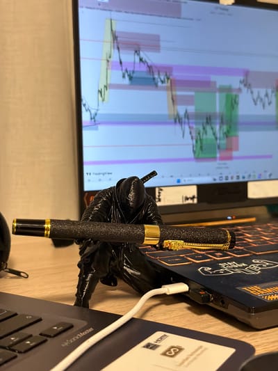 A black, textured fountain pen with gold accents and a skull clip is prominently displayed. The pen is held by a glossy, black gloved hand or mannequin hand. In the background, a computer monitor displays a chart with colorful bars and lines, likely related to financial trading or data analysis. To the right of the pen, there is a laptop with "TUF GAMING" sticker, a keyboard, and a HDMI cable plugged into a port. There is a wooden surface in front of the items. The environment provides no indication to a particular city or location.