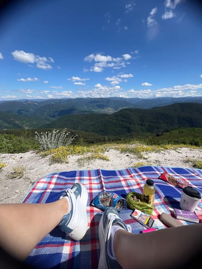 The image shows a first-person perspective of someone sitting on a red, white, and blue checkered blanket at a scenic overlook. The view is of rolling, green, forested mountains under a bright blue sky with scattered white clouds. In the foreground, on the blanket, are various items including a bottle of yellow liquid, a pack of sunflower seeds, a coffee cup, a pack of cigarettes, a pink lighter, and a beige object that appears to be a vape pen. The person is wearing white sneakers with blue accents and white socks. The ground beside the blanket is rocky with sparse yellow flowers and a bush with light-colored, feathery foliage. The location appears to be a natural, elevated area, possibly a mountain or hilltop, offering a panoramic view of a valley and further mountain ranges. There is no indication of it being in the city of Kosovo, Bulgaria.