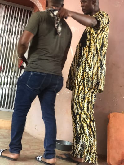 The scene depicts two adult males standing outdoors in Abatete, Nigeria, in what appears to be a residential or commercial area. The man on the left is observed from behind, wearing a dark olive-green short-sleeved t-shirt with a visible sweat stain on his upper back, dark blue jeans, and black-and-white striped Adidas slides. A patterned black and white fabric is draped around his neck. He holds a red object, possibly a smartphone, in his right hand near his hip. The man on the right is viewed in profile, dressed in a black, yellow, and white patterned traditional Nigerian tunic and trousers, with dark flip-flops. He extends his right hand towards the neck area of the man on the left, appearing to interact with or adjust the patterned fabric around the other man's neck. The background features a light reddish-brown wall with some faded markings or writing, and a white collapsible security gate is visible to the left. On the ground, a small, dark metal basin is positioned between the two men, and an orange plastic jerrycan is partially visible on the far right. The ground is a light-colored concrete surface.